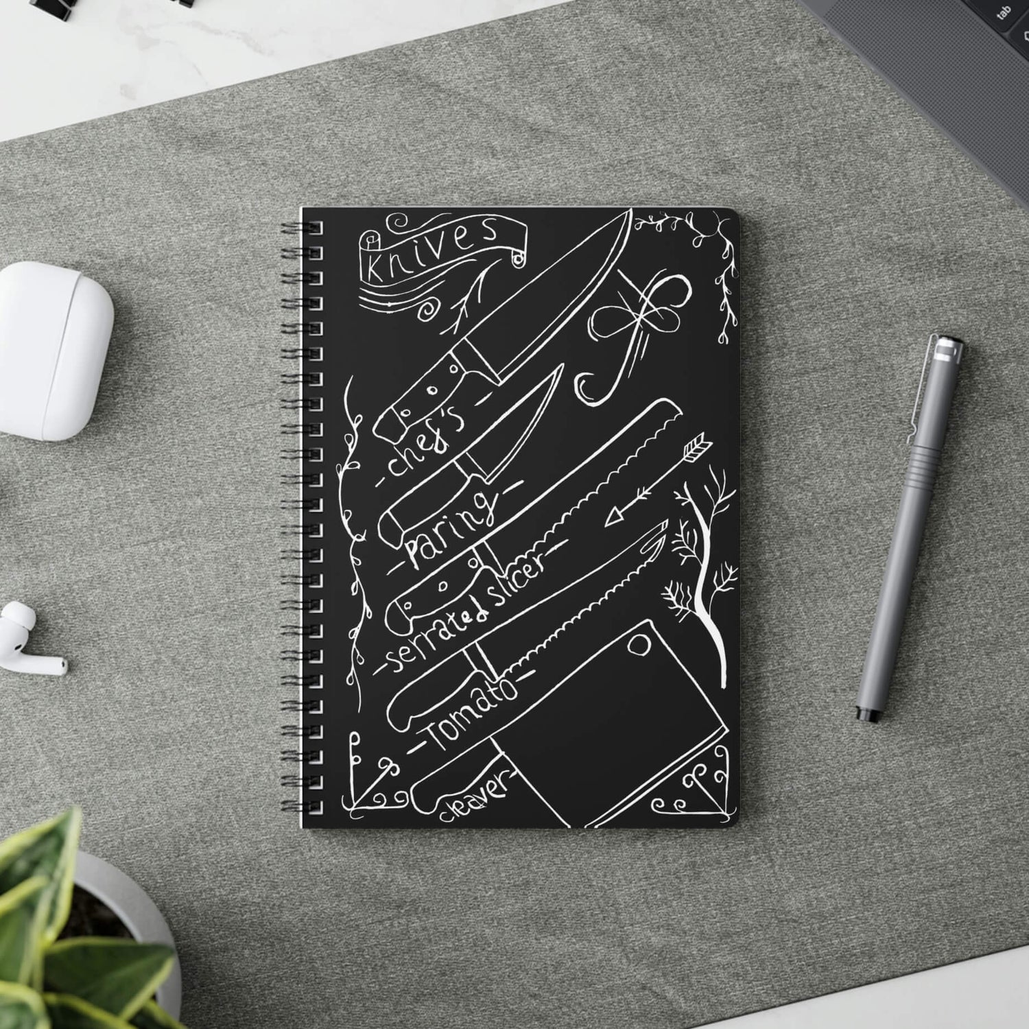 a wire binded notebook on a work top with a digitally edited lino print of kitchen knives